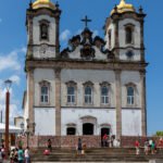 Gradil de ferro ornamentado na fachada da Igreja do Senhor do Bonfim, em Salvador, coberto por centenas de fitinhas coloridas do Bonfim amarradas pelos fiéis, símbolo de pedidos e promessas de devoção popular.