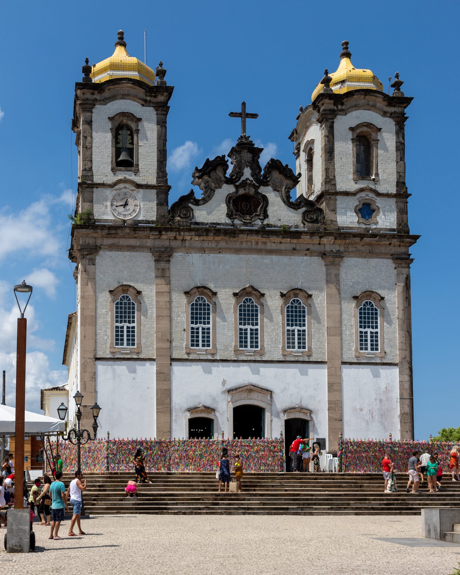 Gradil de ferro ornamentado na fachada da Igreja do Senhor do Bonfim, em Salvador, coberto por centenas de fitinhas coloridas do Bonfim amarradas pelos fiéis, símbolo de pedidos e promessas de devoção popular.
